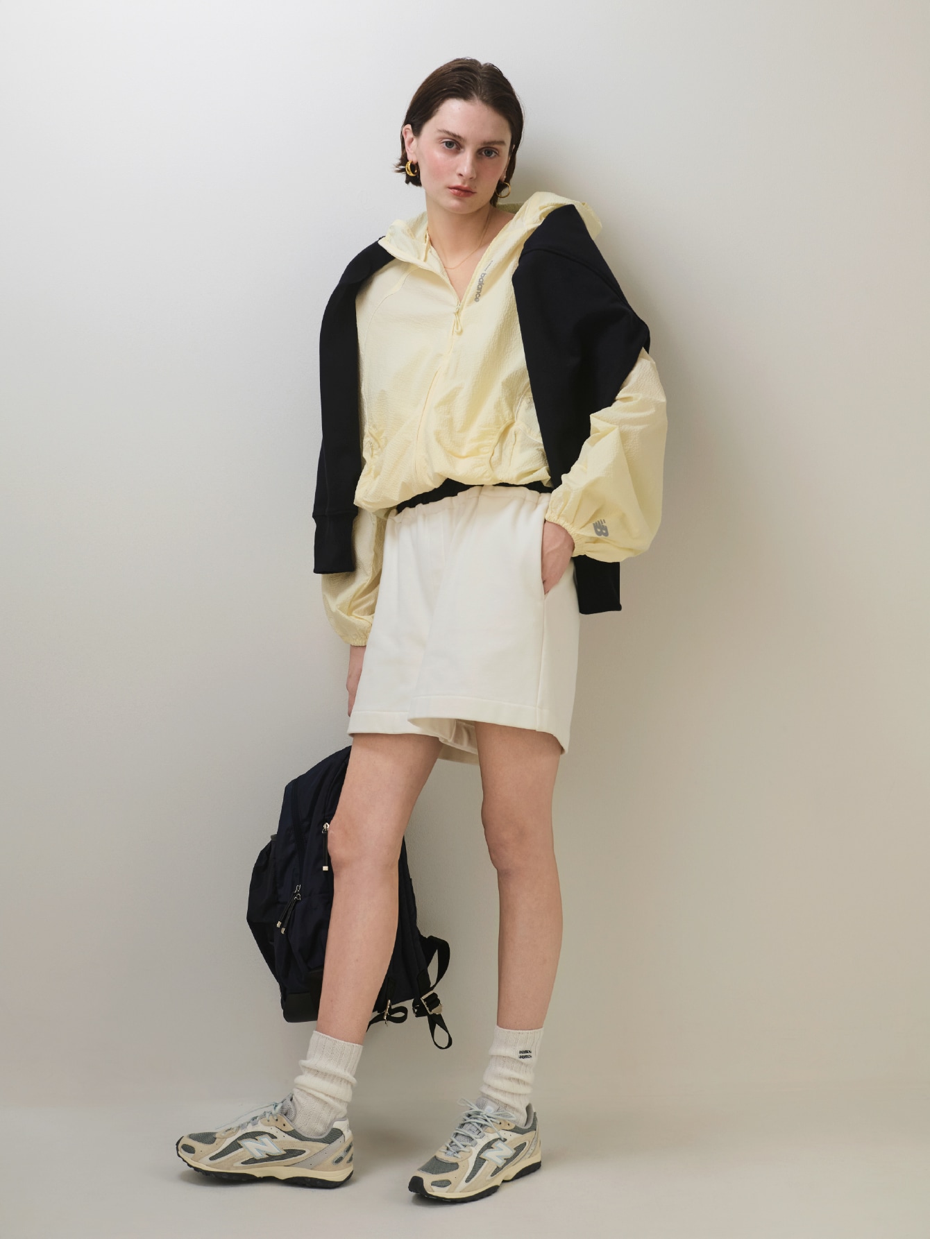 イエローのマルチファンクションジャケット・ネイビーのハーフジップスウェット【セットアップ対応】・白のレイヤードライクスウェットショートパンツ【セットアップ対応】・ネイビーのecoレザーミックスバックパック・アイボリーのレギュラーソックス・グレーのU204Lを合わせたコーディネート