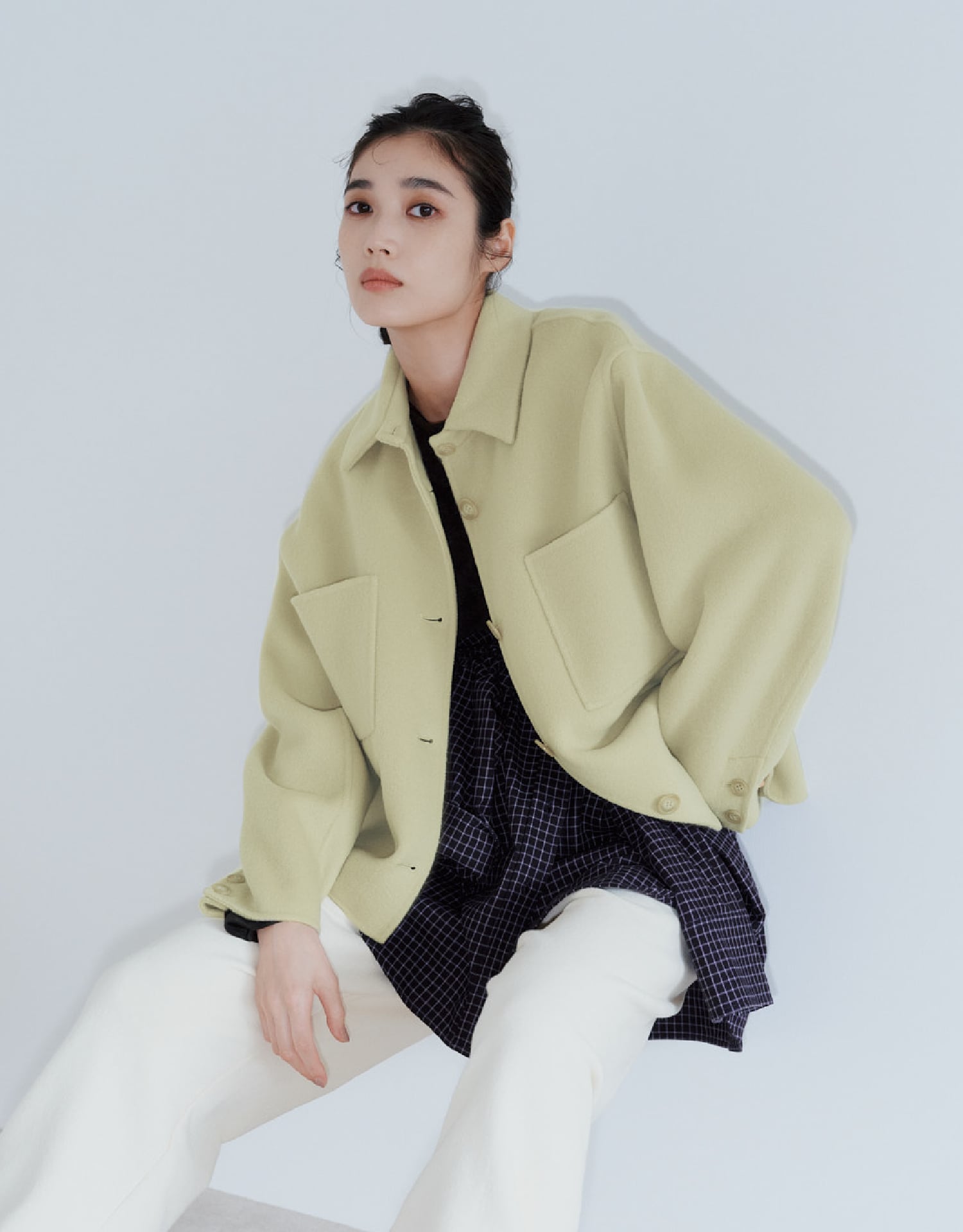 オリーブ色のジャケットとネイビーのチェックシャツ、アイボリーのパンツを着用して壁に持たれる女性