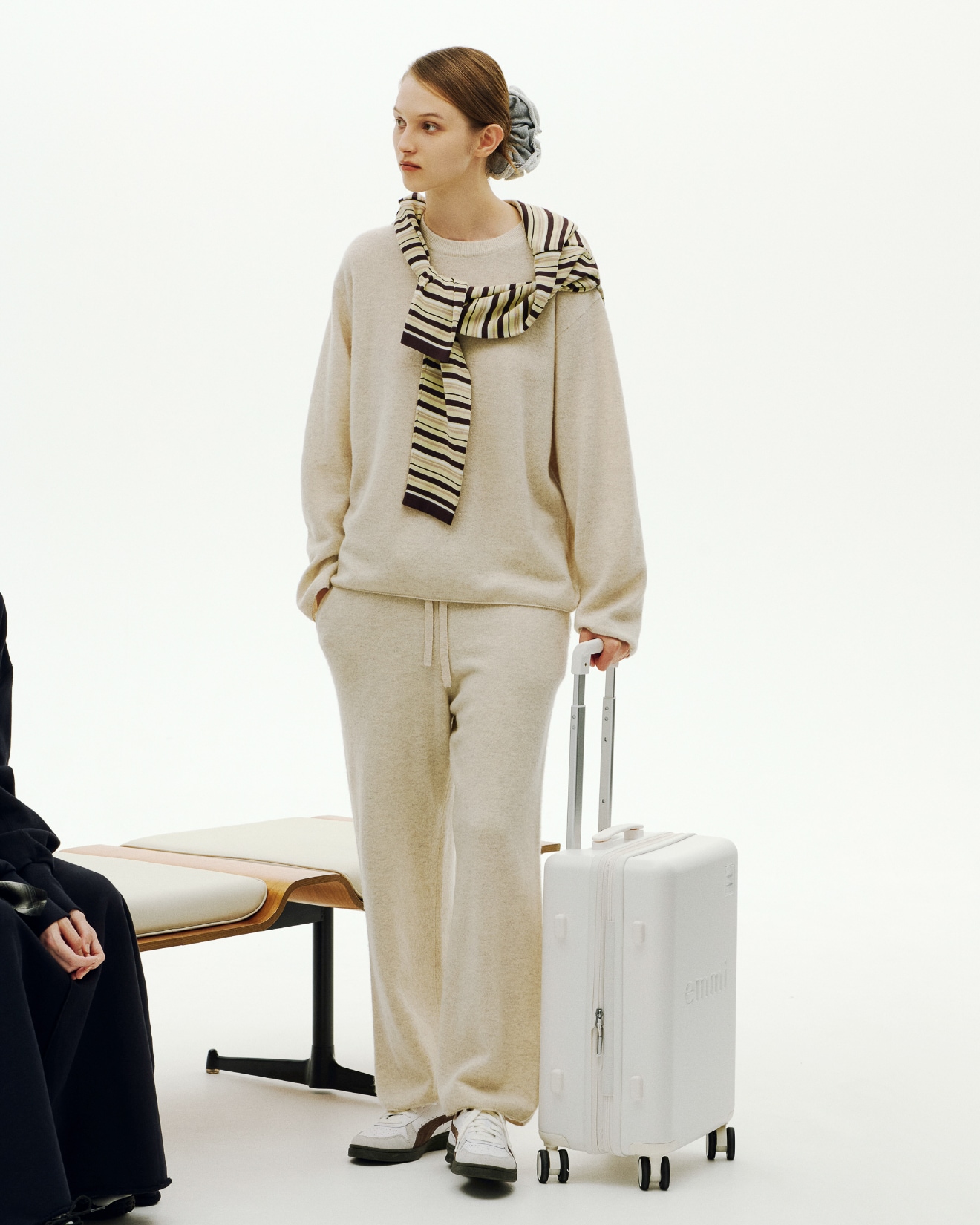 ライトベージュのラムウールオーバーニットトップスとパンツ・イエローの ボーダーニットとグレーのスウェットシュシュ・Indoor（シューズ）を着用し、Samsonite スピナー55 のハンドルに手をかける女性のビジュアル