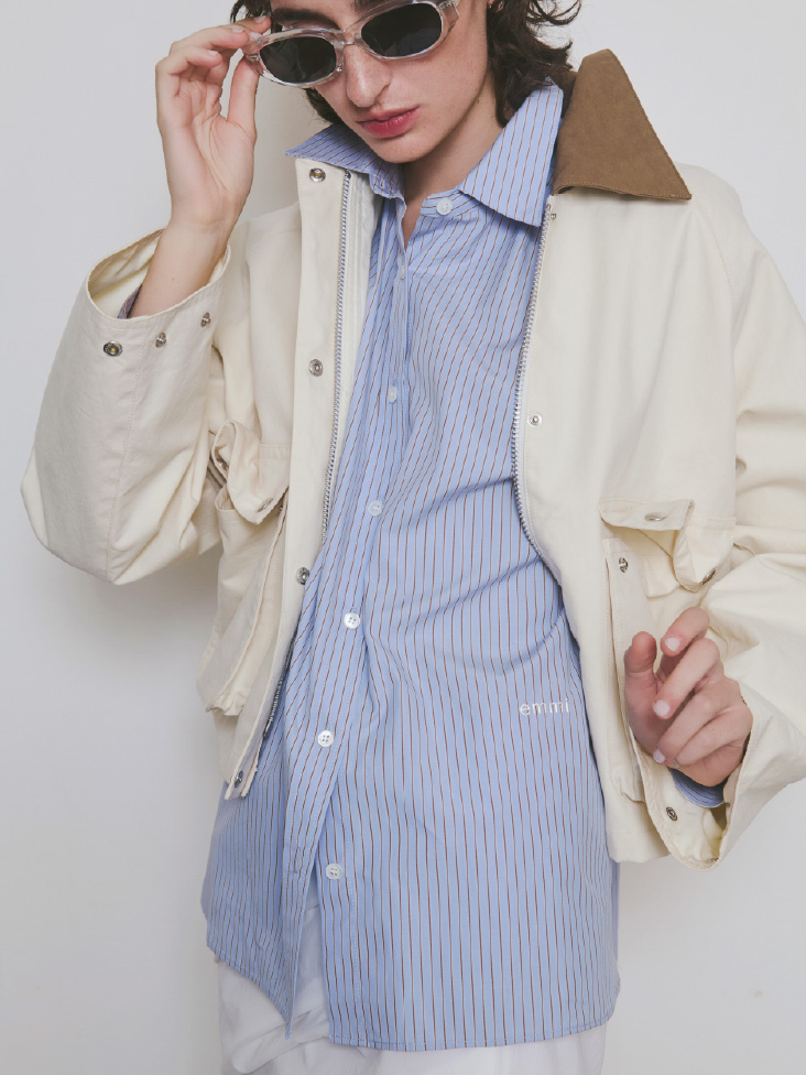 アイボリー色のショートハンティングジャケット、ブルー色のシャツ、オーバルキャットアイフレームサングラスを着用したモデル画像