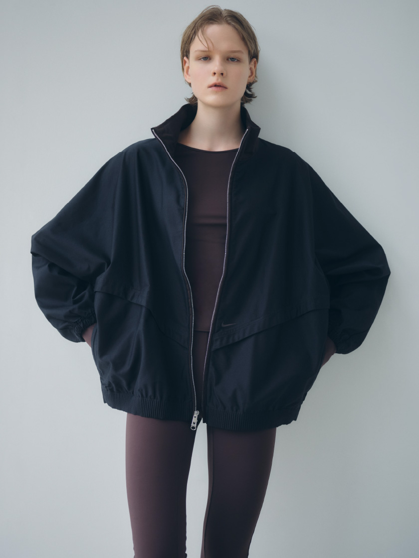ブラック色のOversized Windrunner Twill Jacketとダークブラウンのサステナレッチカプリレギンスを着用した女性の後ろ姿